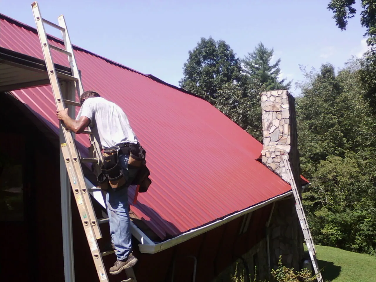 Roofer on a ladder beside a red metal roof and stone chimney — Soffit & Fascia Repair by Roofing Brothers in Wauconda